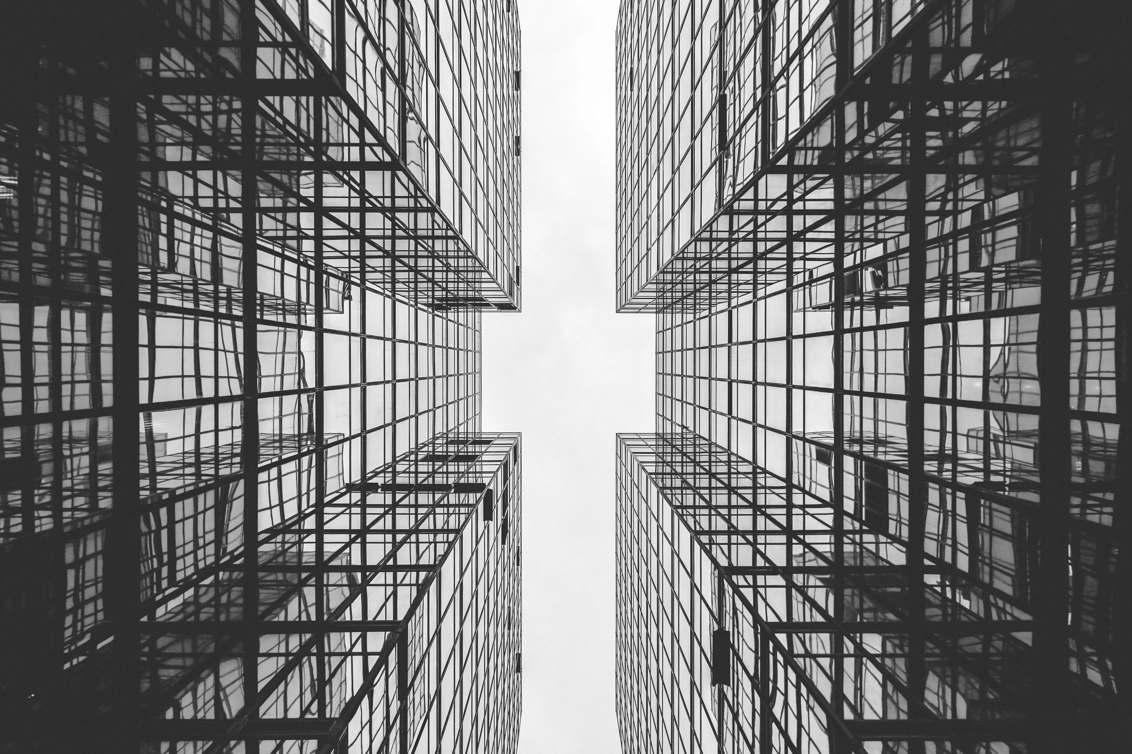 Monochrome photograph looking up between two mirrored glass elevations, their window grids meeting at a narrow strip of sky