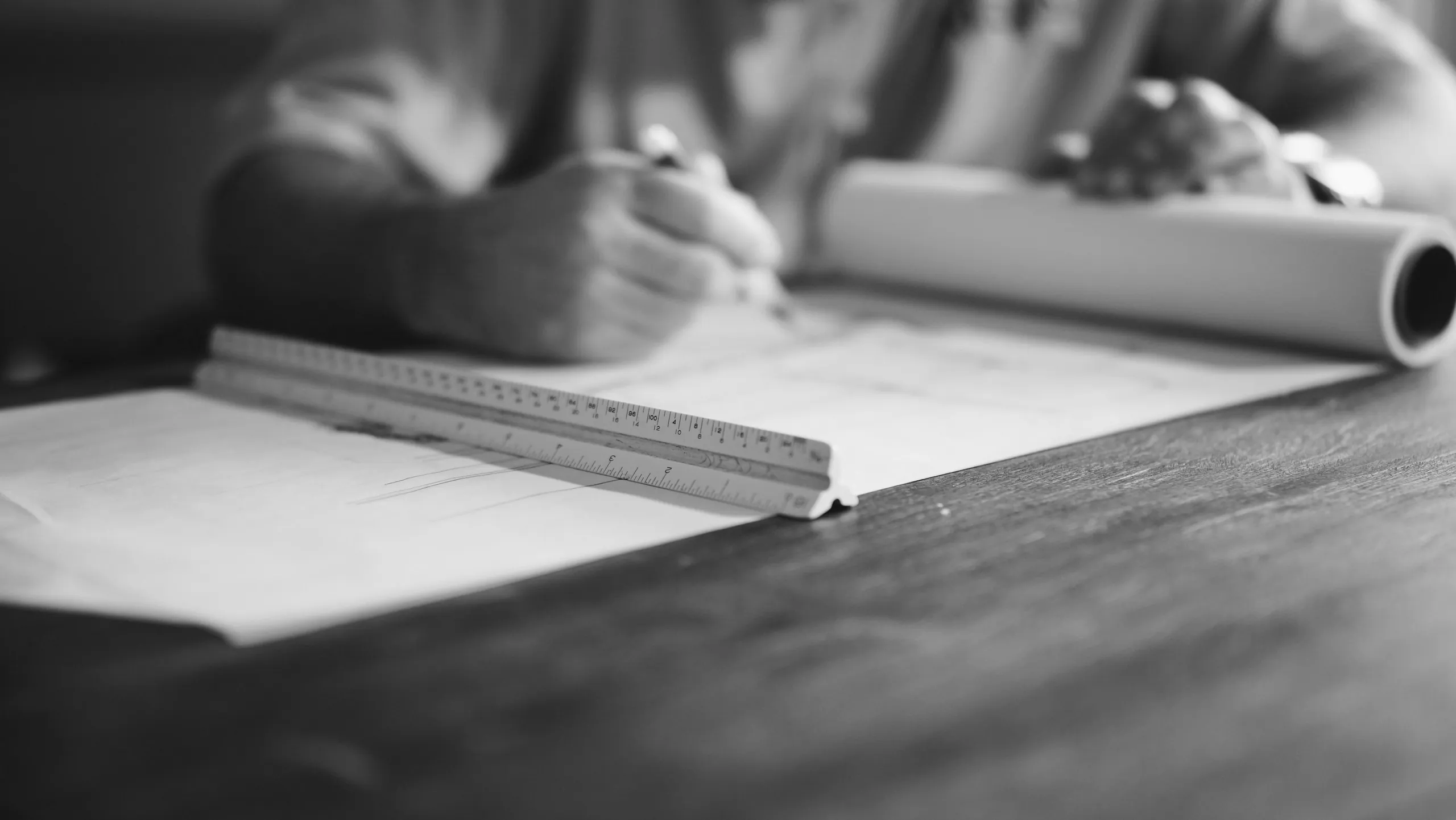Monochrome close photograph of hands drawing on paper at a wooden table, with a triangular scale rule and a rolled drawing alongside