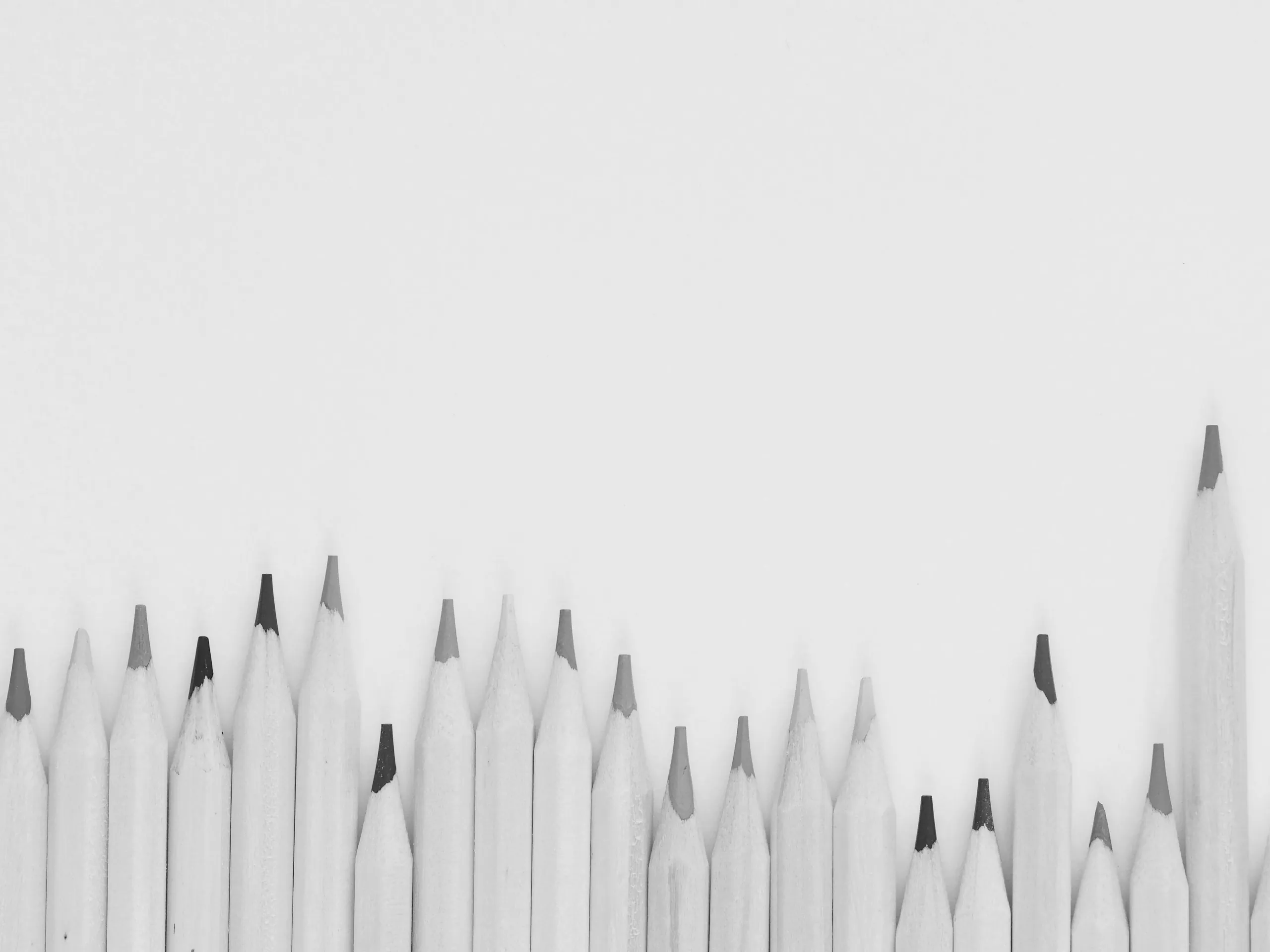 Monochrome photograph of a row of sharpened pencils stood upright against a plain pale background, their graphite points at uneven heights