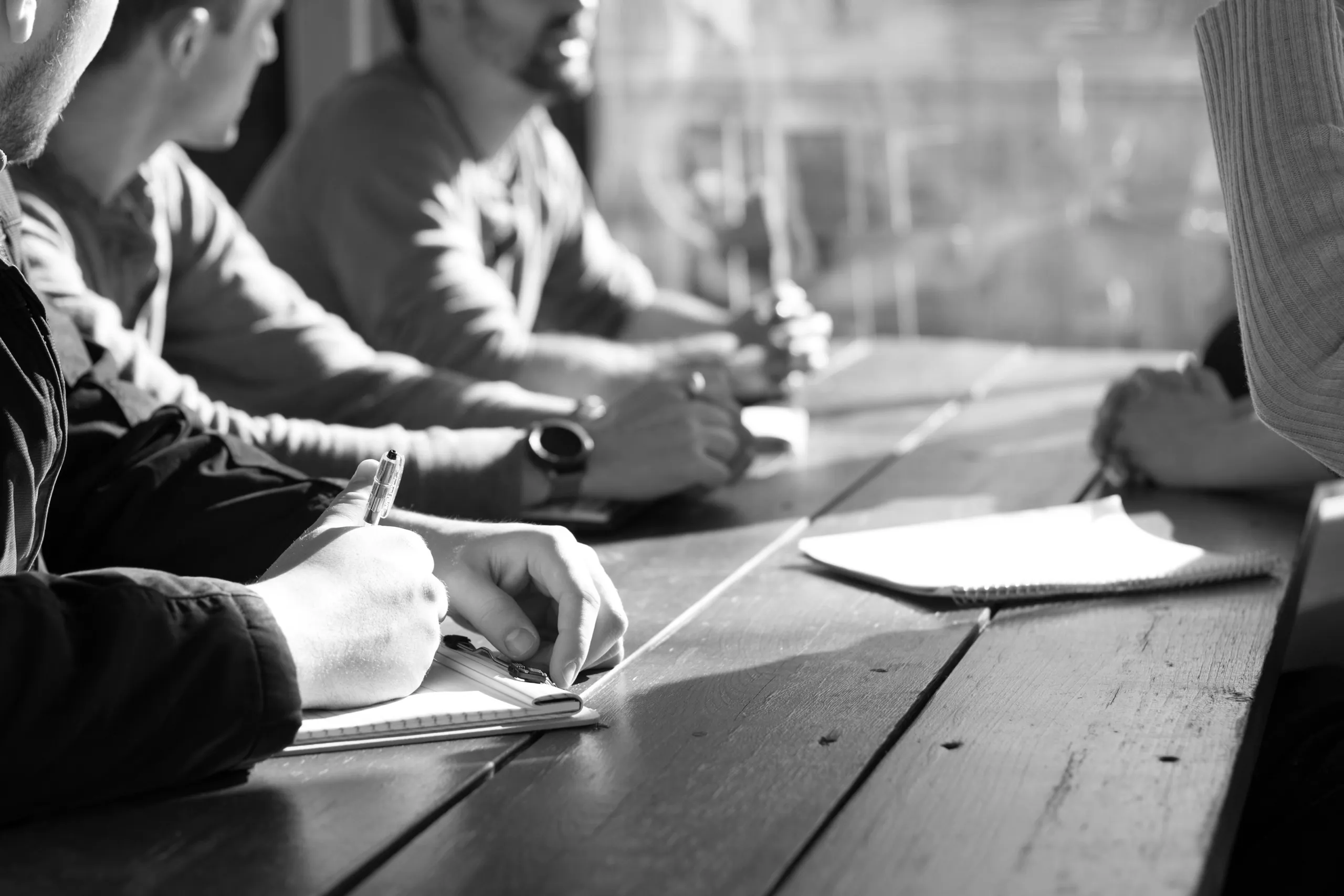 Monochrome photograph of four people around a long timber table, writing in notebooks in raking side light