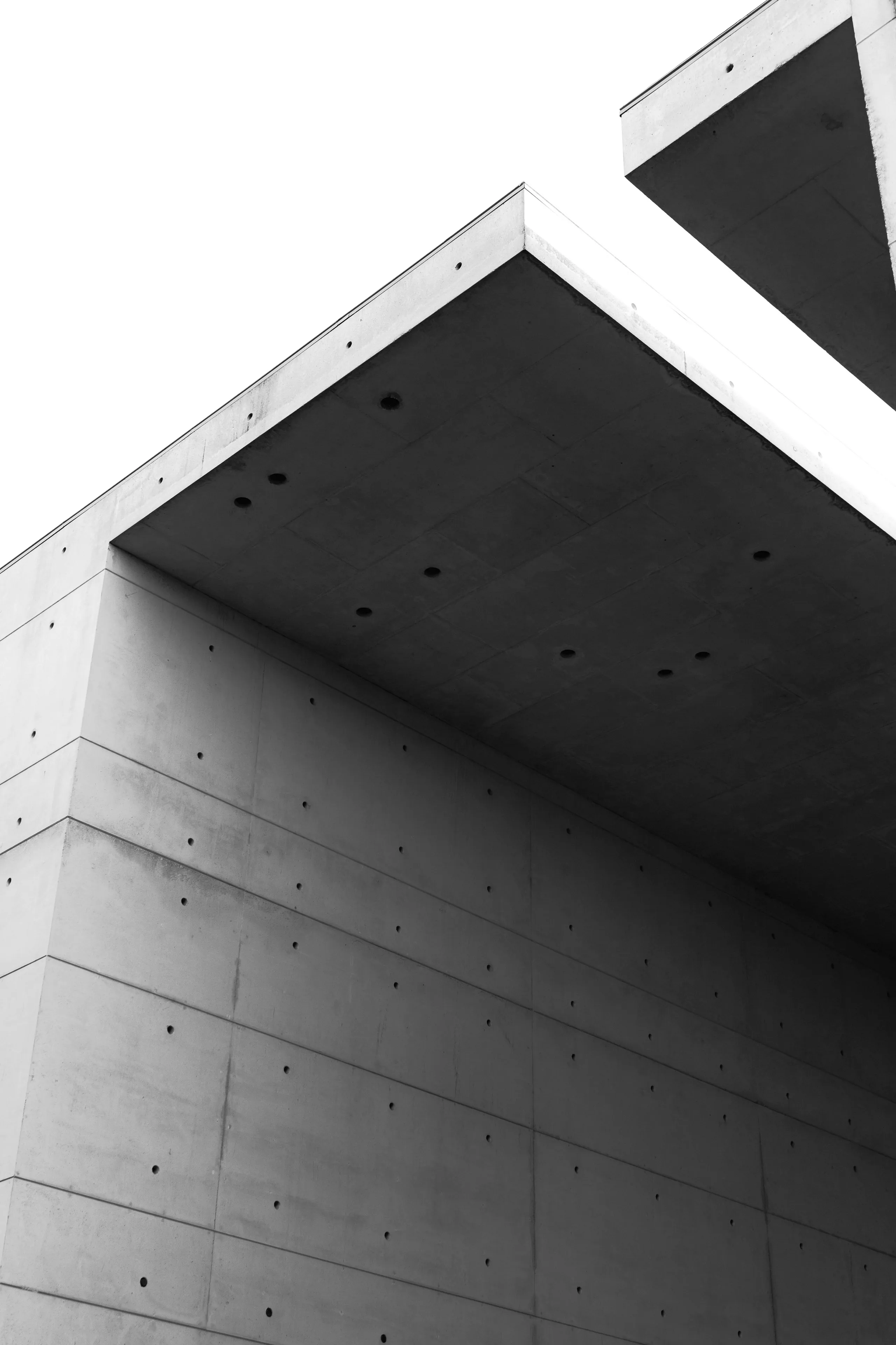 Monochrome photograph looking up at a concrete building corner: a deep soffit oversailing a wall of panels marked with regular tie holes