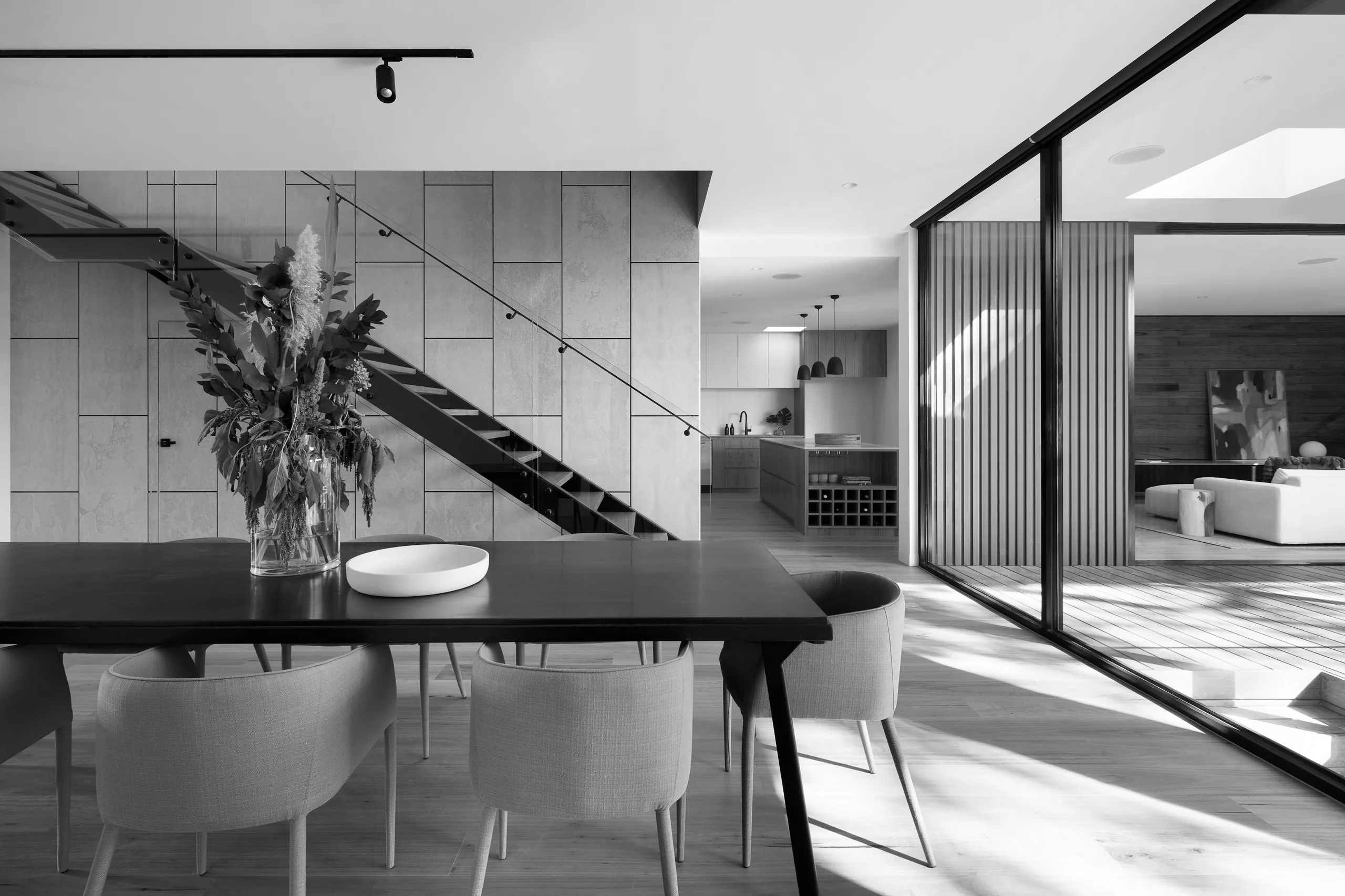 Monochrome photograph of an interior: a black steel open-tread stair against a panelled wall, a dining table in the foreground and a glazed screen to the right