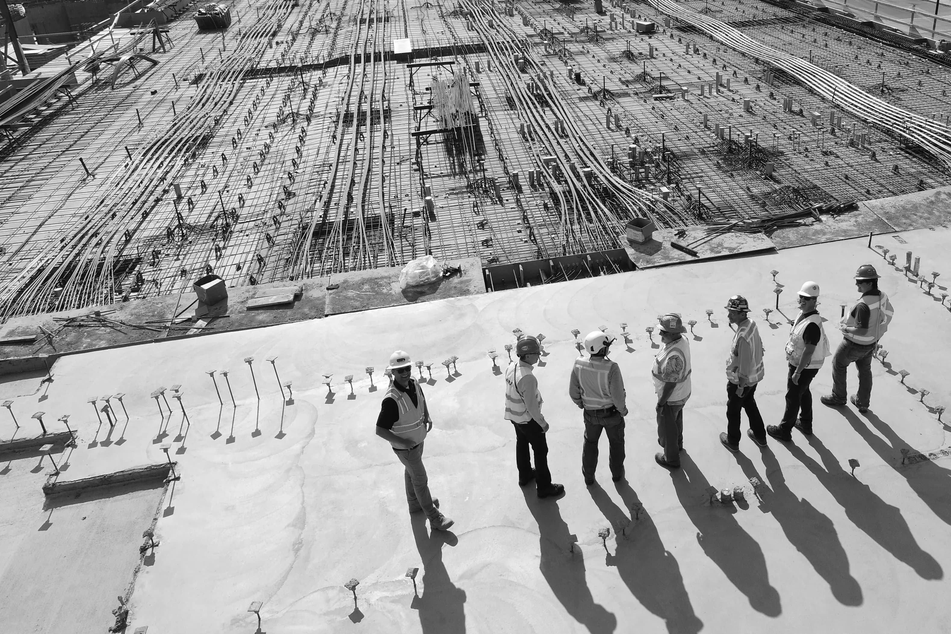 Monochrome photograph looking down on a construction deck: a poured concrete slab with a crew standing in a line, and a dense mat of reinforcement bar beyond