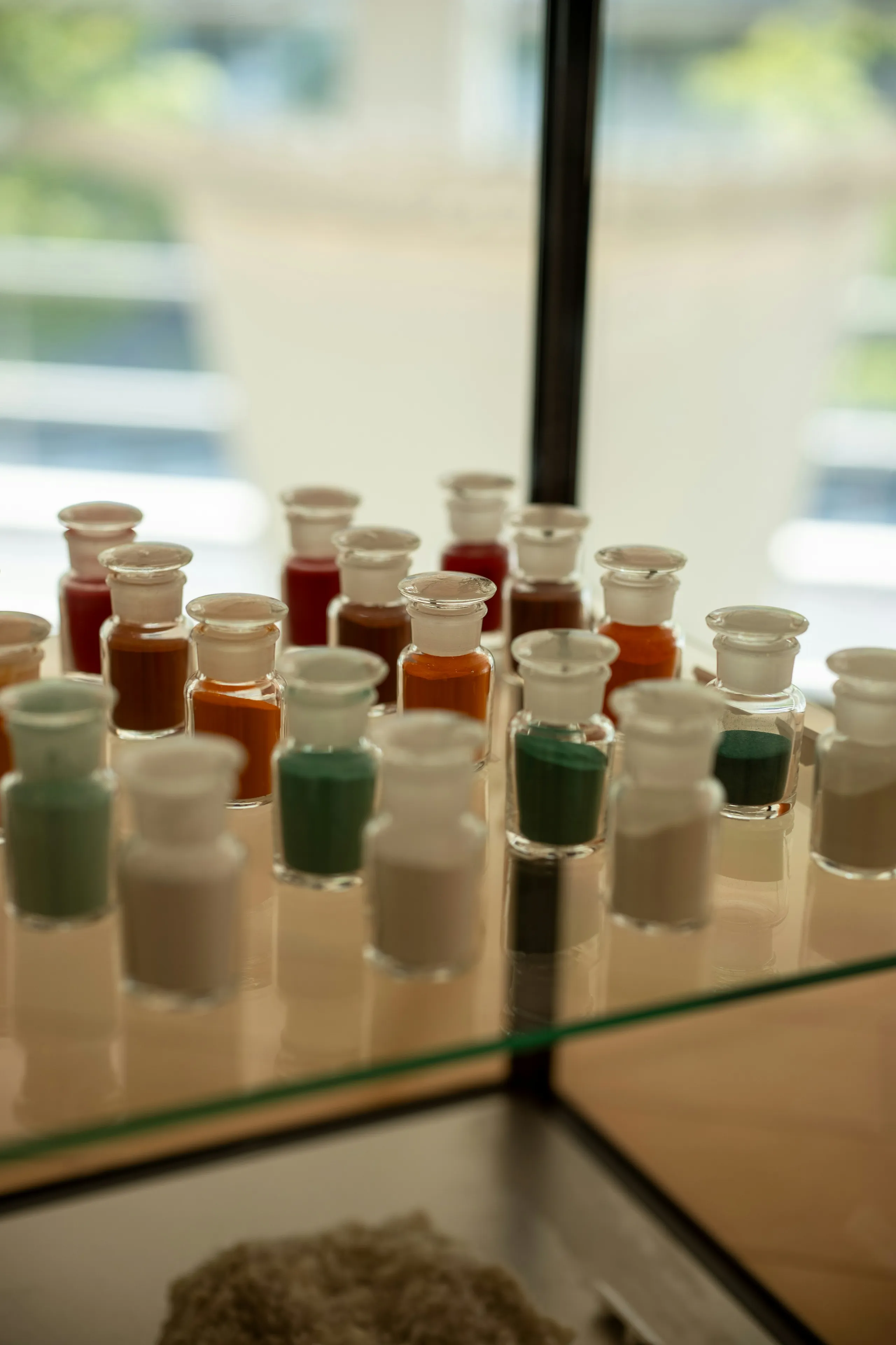 Rows of small glass apothecary bottles filled with dry pigment — reds, ochres, greens and chalk white — on a glass shelf