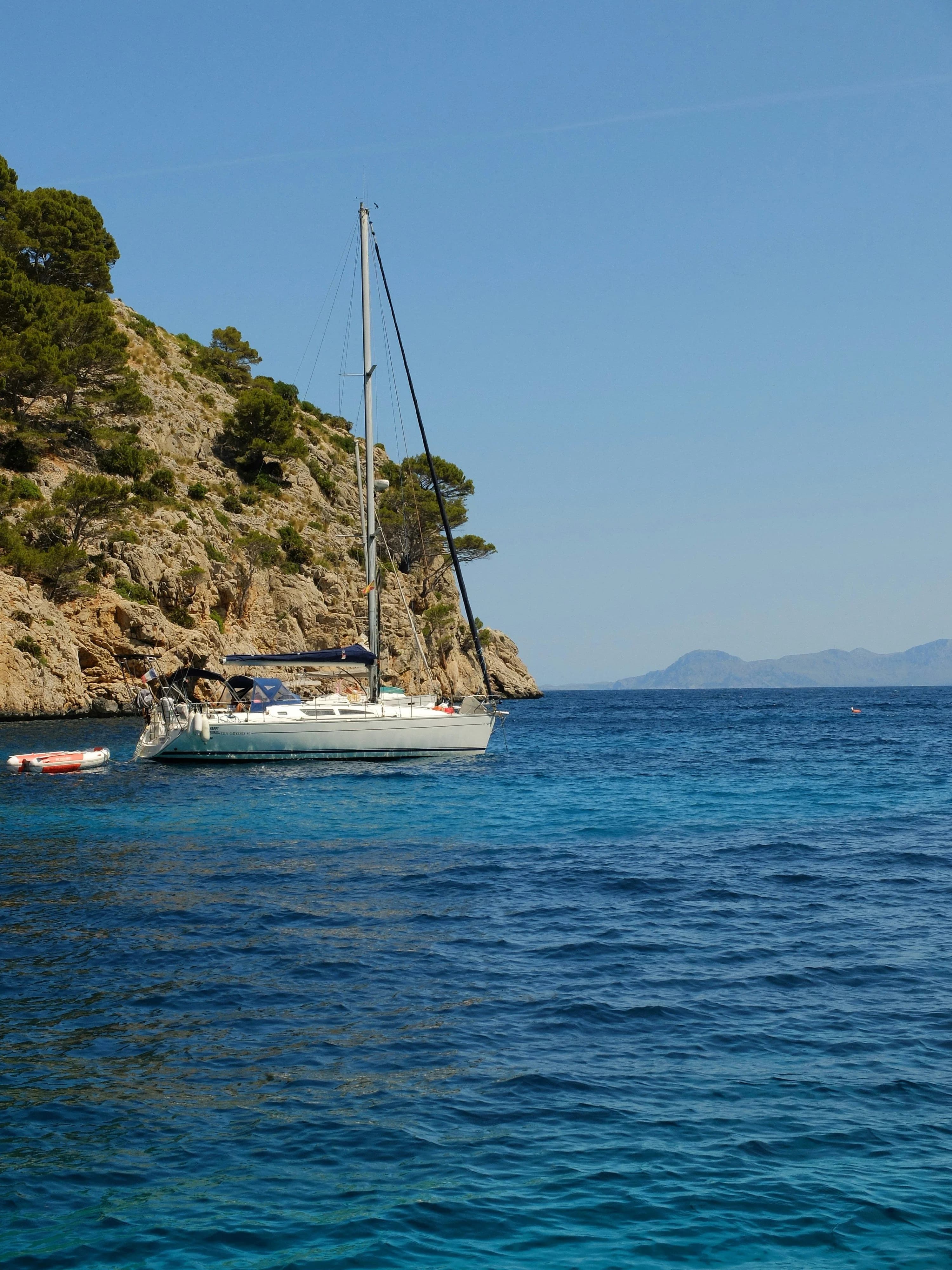 A white sloop lying to her anchor in a pine-fringed cove, her tender astern, clear turquoise water beneath her