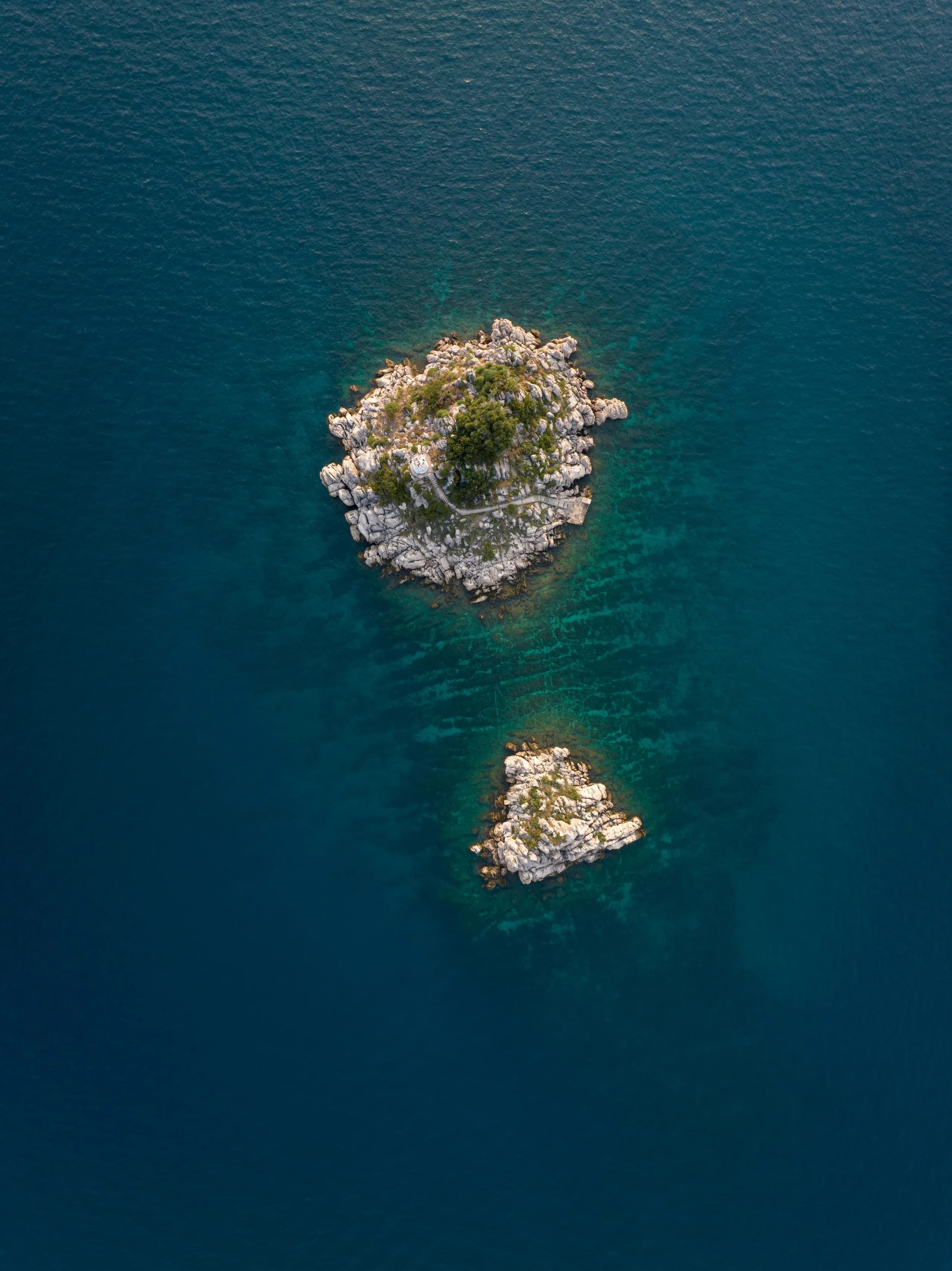 Two bare rock islets alone in deep teal water, seen straight down from the air