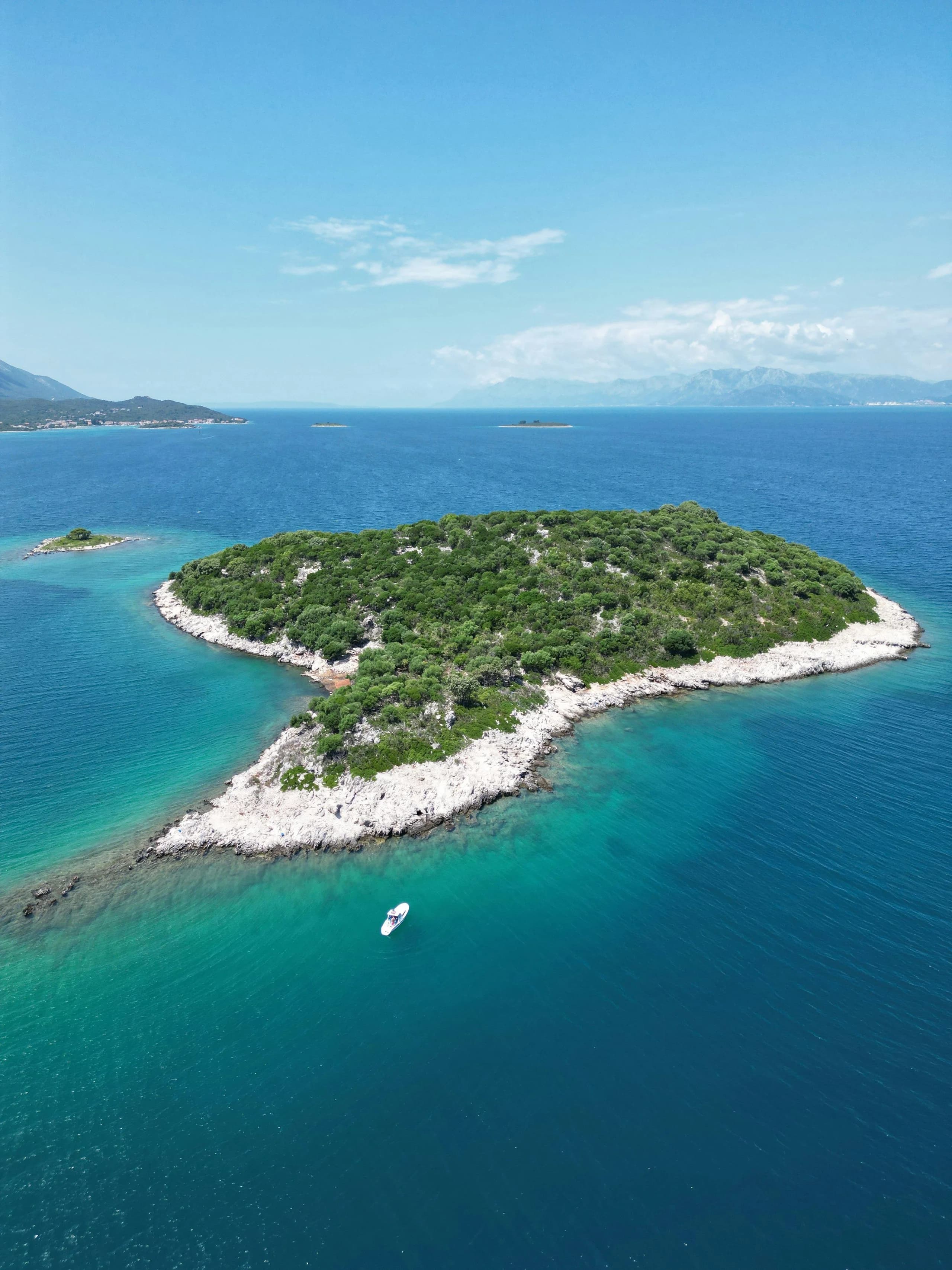 Aerial view of a low wooded island ringed with white rock and turquoise shallows, the mainland mountains beyond